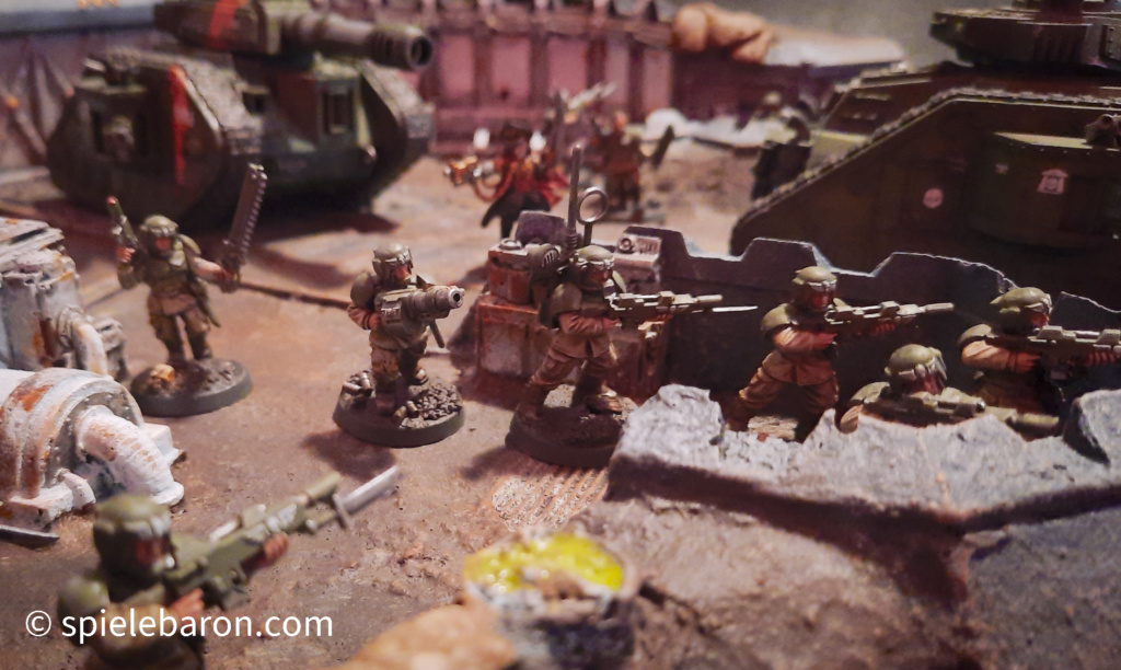 40k Showcase Foto: Astra Militarum, bemalte Imperial Outpost Spielplatte von Forge World, darauf Miniaturen von Leman Russ Panzern und Infanterie Soldaten, gehobener Standard