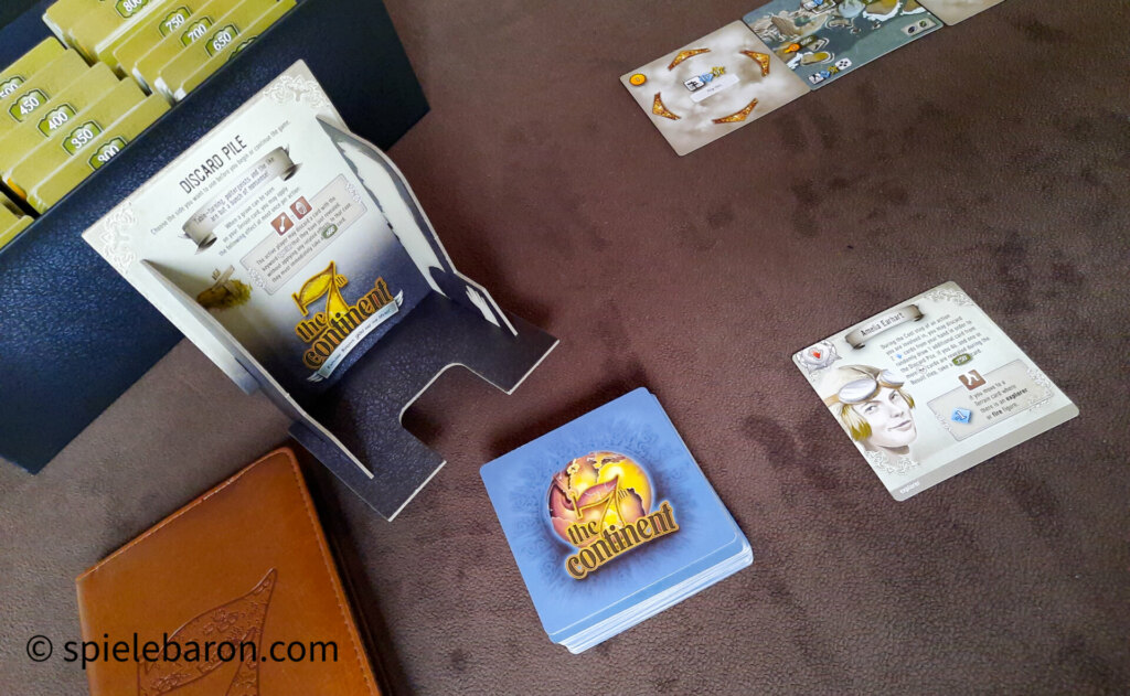 Foto der Spielkomponenten bei Spielaufbau für eine Partie The 7th Continent: Kartenbox, Behälter für Ablagestapel, Journal für die Sicherung unserer Tagebucheinträge, Actiondeck und Startkarte für Amelia Earhart